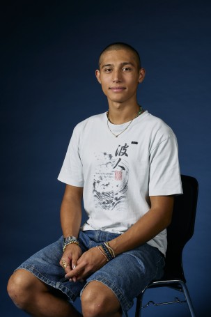 C'est la photo portrait de Keanu bergonzoli, c'est l'apprenti technisceniste du théâtre. Il a les cheveux rasé, il porte un bermuda jeans, un t-shirt blanc. Il a la vingtaine il est assis sur une chaise et il croise les mains sur ses jambes.