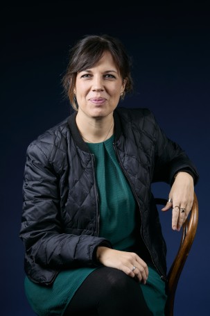 C'est la photo portrait de Aurélie Menaldo. Responsable ecoles et chargée de production. C'est une femme blanche sur la quarantaine.Elle a les cheveux attaché et une frange rideau brune. Elle porte une veste noire et une robe verte.