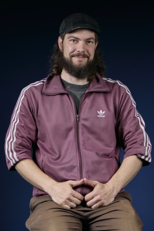C'est la photo portrait de Daniel Emery, régisseur technique. Homme blanc sur la trentaine. Il porte un chapeau il a les cheveux et une barbe brune. Il porte une veste adidas bordeau clair.