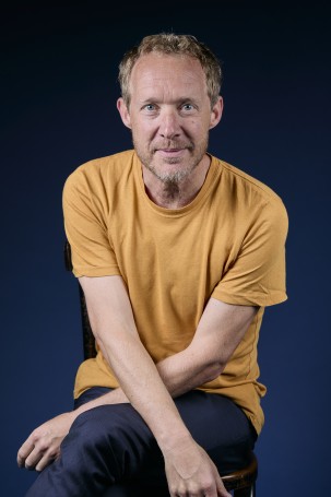 C'est la photo du directeur du théâtre, Eric Devanthéry. Homme, blanc, la cinquantaine, le teint blond, il porte un t-shirt jaune, il regarde la caméra il croise les bras sur ses jambes.