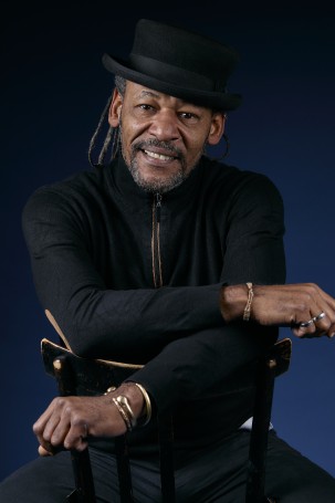 Il s'agit d'une photo portrait du directeur technique du théâtre Julien Okoué Metogo. Homme noir la cinquantaine, porte un pull et un chapeau noir. Il a des dreds et une petite barbe.Il est assis au contraire sur une chaise comme sur un cheval. Il regarde la caméra, il sourit.