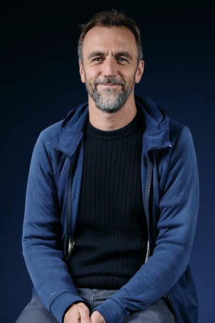 C'est la photo de Marc Erwan Le Roux. L'administrateur du théâtre. C'est un homme blanc sur la cinquantaine.Il porte un t-shirt noir et une veste bleu. Il sourit en regardant la caméra. Il a une barbe poiuvre et sel.