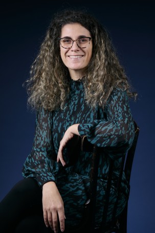 C'est la photo de Marilù Calì, elle s'occupe de la communication digitale, de l'inclusion et des relations publiques. C'est une femme blanche sur la quarantaine. Elle a les cheveux blonds longs et bouclés. Elle porte une robe verte et noire. Elle est asise de profil et elle regarde la caméra.