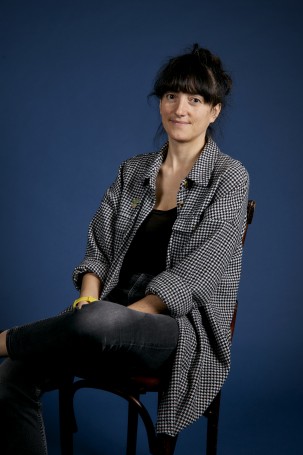 Une personne assise sur une chaise, les cheveux bruns, une frange. Elle porte une veste à carrés noir et blanc. Elle croise un peu les jambes, elle regarde la caméra.