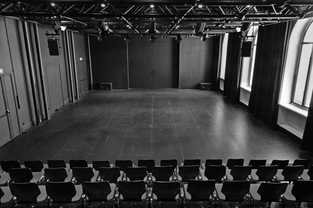 Une photo de la salle du haut depuis le public. On voit la scène vide, une boite noire et les chaises des premieres rangées.