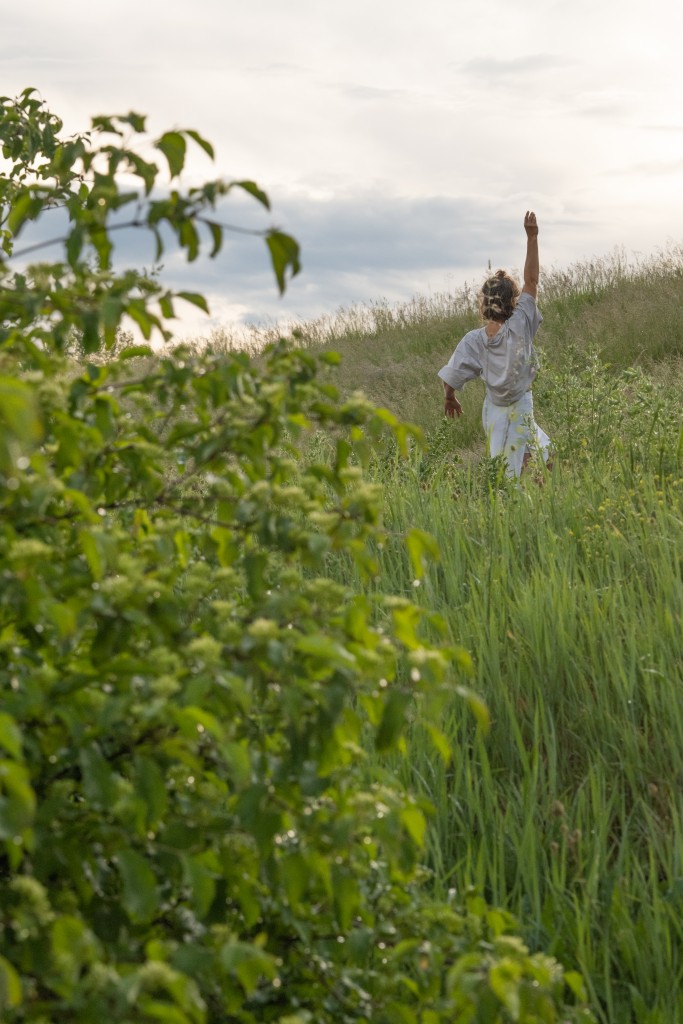 Une personne de dos en train de danser. On voit qu'elle lève le bras. Elle est au milieu des hauts herbes.