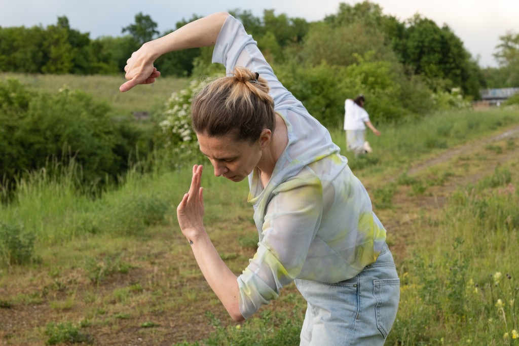Deux personnes en train de danser. Une plus proche de la camèra, l'autre plus loin. les deux sont comme dans un champs ou un milieu vegetalisé.