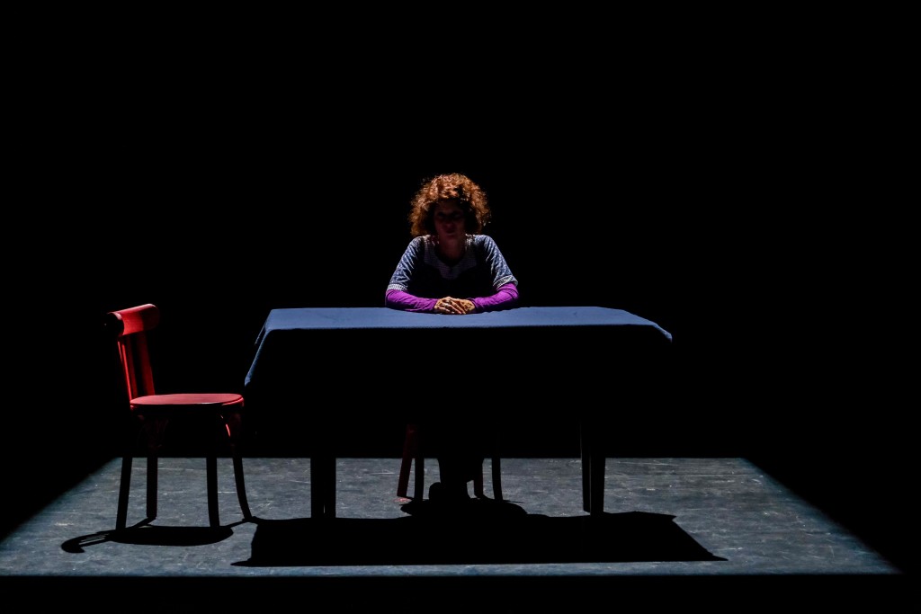 Une personne assise derrière un table. On ne voit pas le visage de la personne car la photo est assez sombre.