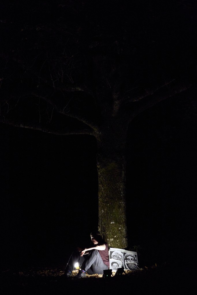 C'est une photo très sombre, prise à la tombée de la nuit. Françoise Boillat est assise au pied de l'arbre, il y a de petites lumière qui illuminenet un peu. On voit des photos noir et blanc contre l'arbre.