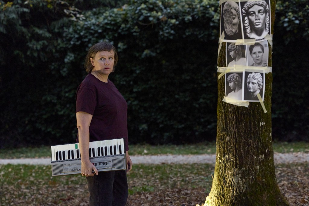 C'est une photo de Françoise Boillat elle porte un petit piano sous le bras. Elle est à côtés du tronc d'un arbre sur lequel sont collées des photos noir et blanc.