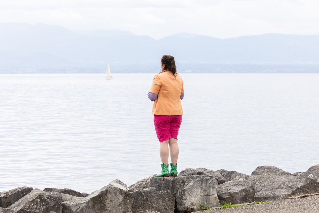 Une personne débout face au lac, elle tourne le dos à la caméra. Elle porte des vetements très colorés.