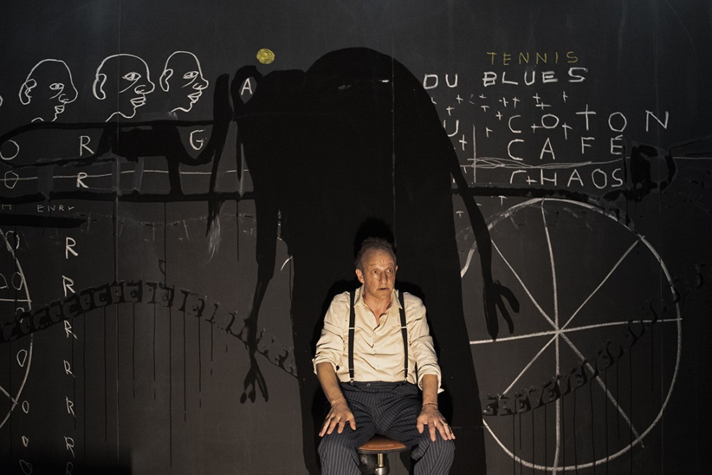 Une personne assise avec l'air appeuré, elle regarde devant. Elle est assise devant un tableau noir et sur le tableau noir ce n'est pas son ombre qui est projettée mais celle d'un monstre.