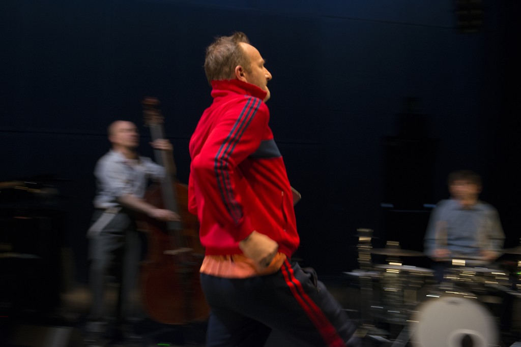 Une personne en training rouge en train de courir. On voit à peine son visage. Derriere lui des personnes en train de jouer de la musique mais l'image de fond est floue.