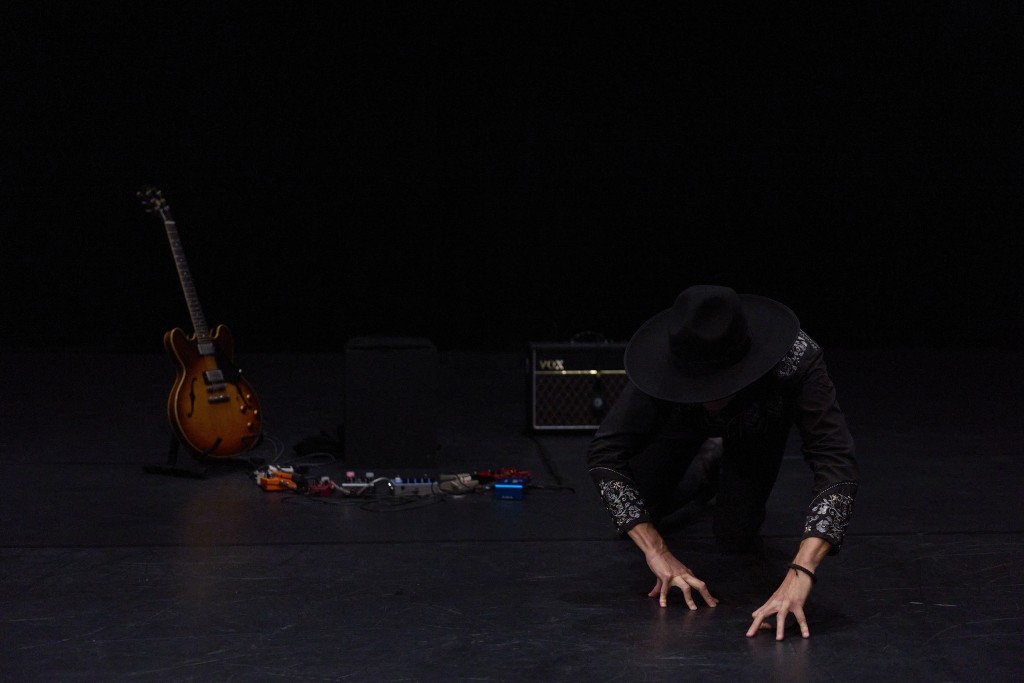 Nicolas Cantillon par terre en train de mimer des gestes avec ses mains. La photo est un peu sombre on voit une guitarre posée sur la droite de la photo.