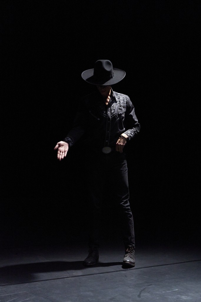 C'est la silhouette de Nicolas Cantillon habillé en noir avec un chapeau de cow boy. Une main sur la ceinture, l'autre ouverte sur le devant. On ne voit pas son visage. La photo est très sombre.