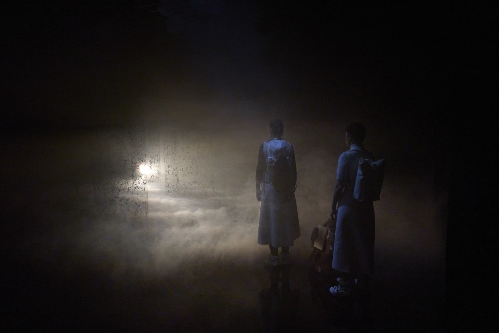 Deux silhouettes habillées en blanc, dos caméra, elles marchent vers une source lumineuse. Il y a du brouillard.