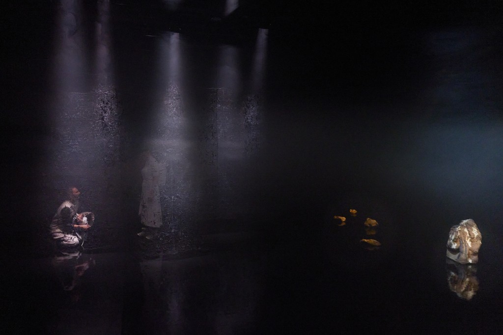 C'est une photo très sombre on aperçoit à peine les silhouettes d'Alexandra Tiedemann et Rachel Gordy, comme derrière une toile. A droite de la photo posé par terre un objet lumineux.