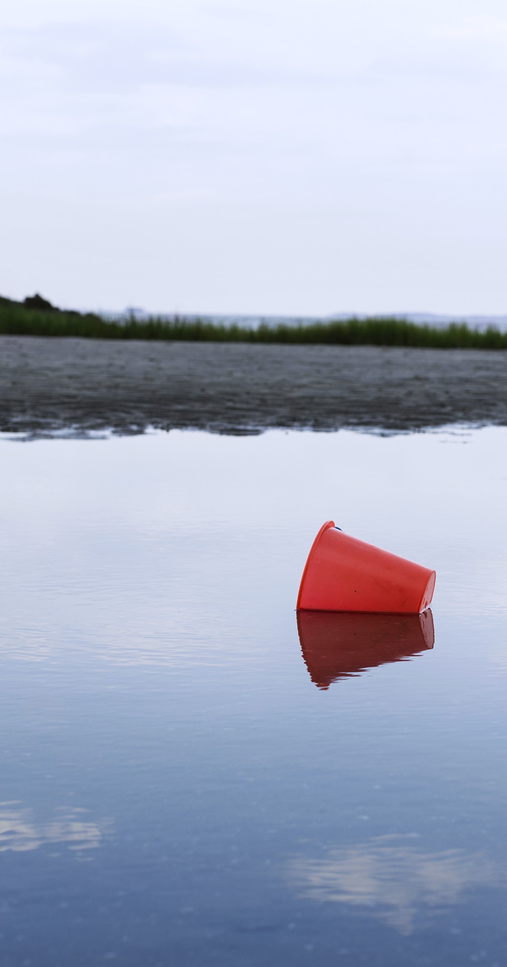 C'est la photo d'un seau rouge oublié dans un etang d'eau on ne sait pas trop si c'est une flaque, un lac, une ribiere, mais l'eau n'est pas très haute. La fin de l'eau se confond avec le debut de la terre et le ciel.