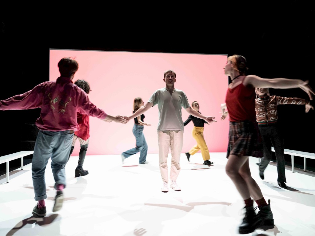 Un groupe de jeunes entre 25 et 30 ans en train de danser, le sol il est blanc, sur le fond une toile fucsia clair. Au côtés des bancs blancs.