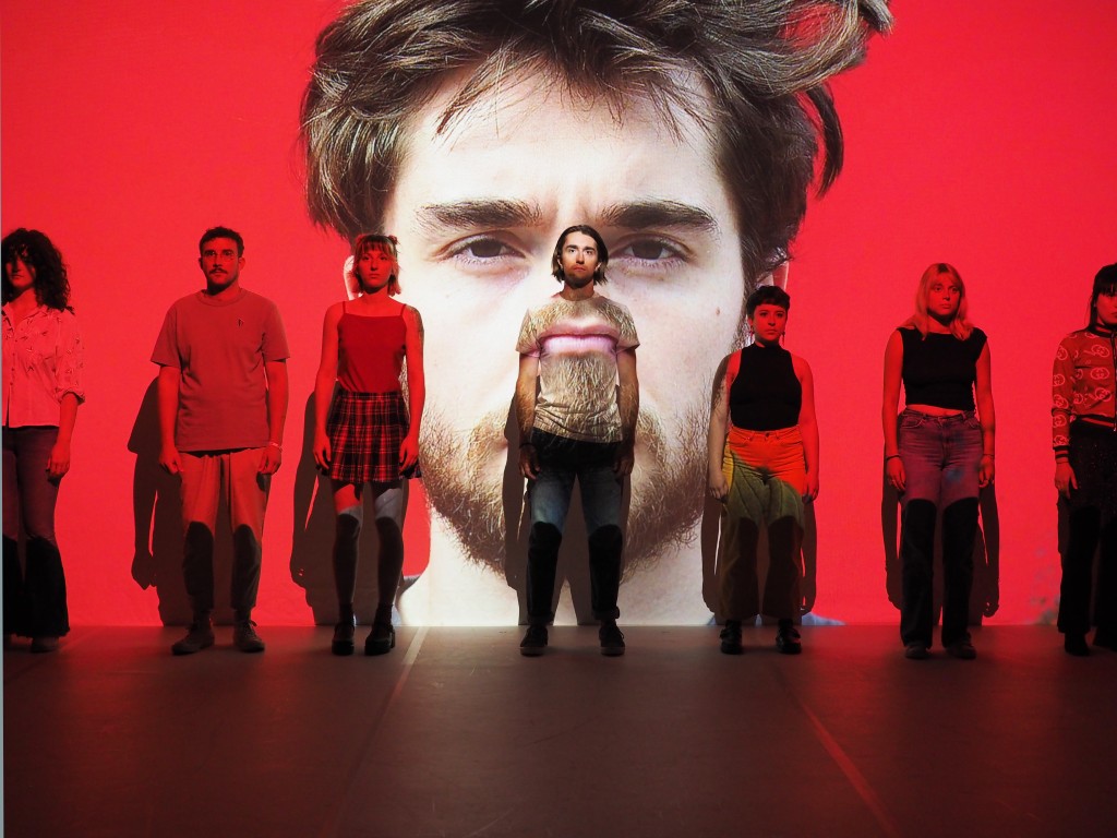 Dans une lumiere rouge, des jeunes appuyés contre une toile sur laquelle est projetté la photo d'un autre jeune.