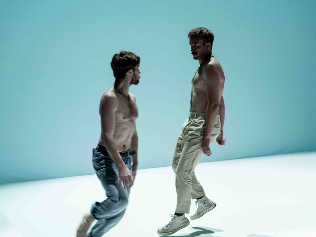 Deux personnes en train de danser. L'une en face de l'autre. Tors nus, sur un plateau blanc.