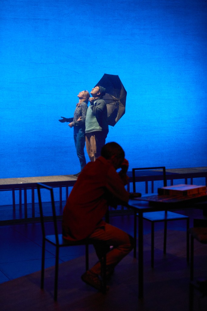 En arriere plan un couple en train de se promener, le couple regarde vers le ciel, le garçon tient un parapluie. Le couple est comme sur un praticable derriere une toile transparente. En premier plan le papa assis sur une chase, très pensif, on le voit à peine appuyer la main sur son front.