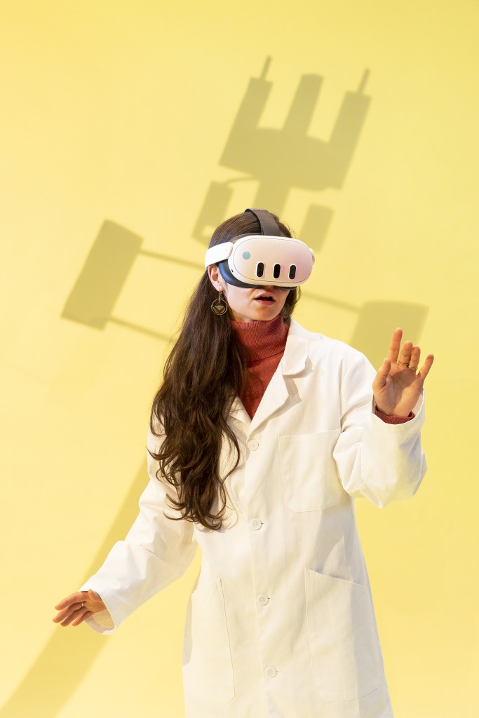 Une personne en blouse blanche avec un casque de realité virtuelle. La personne a les cheveux longs.