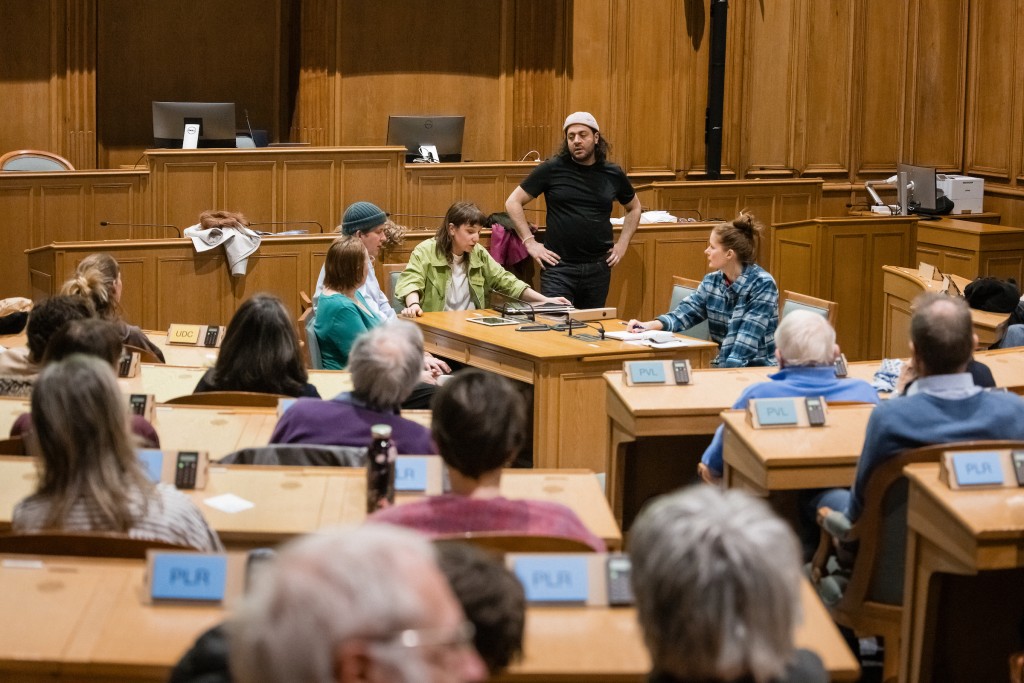 Des personnes assises dans la salle du Grand Conseil, une personne debout en train de parler les mains aux hanches.