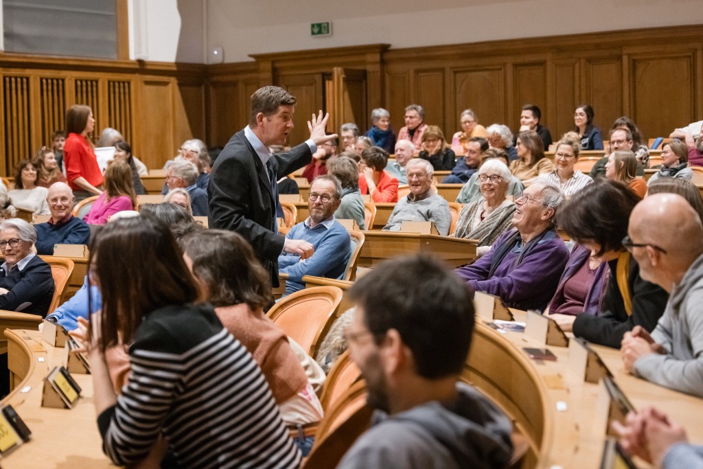 Des personnes assises dans la salle du Grand Conseil, une personne debout en tran de parler.