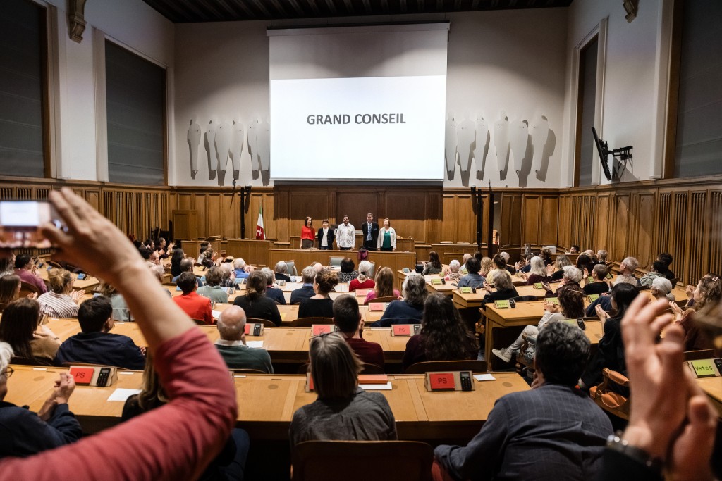 Des personnes assises dans la salles du Grand Conseil, on voit projeté sur un écran au fond le mot : Grand Conseil.