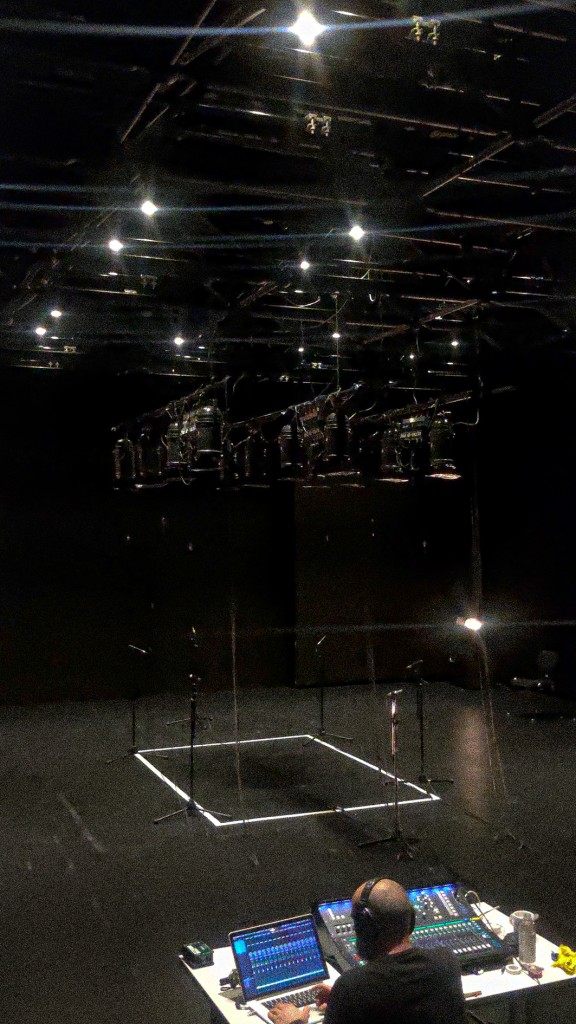 On voit une personne derrière une table de régie lumières. la photo est prise depuzis le public. Sur la scène un carré blanc fait avec du scotch autour duquel il y a des micros sur pieds. En haut plein de projecteurs lumières.