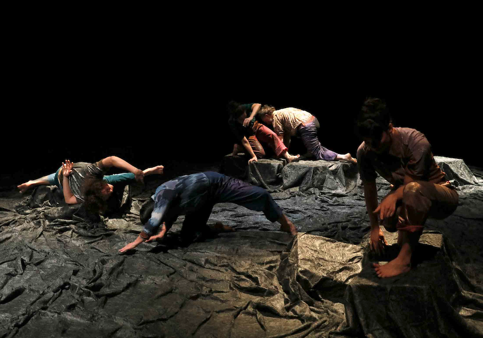 5 personnes en train de danser. certaines se trouvent sur des especes de pierres. Le sol et les pierres sont couvertes comme d'un tissu qui donne l'impression d'un liquide ou du bitume. La photo est sombre