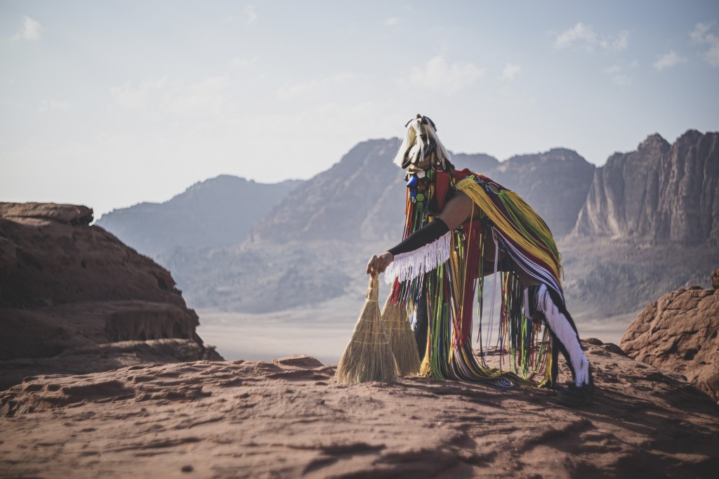 Une personnes habillée avec un costume plein de fils de tissu colorées. Elle est accroupie sur deux balayettes comme en train de ramasser des choses avec les deux balayettes. Derrière cette personne un paysage de montagne et un lac. Le paysage a l'air aride.