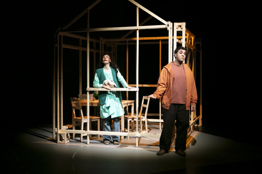 Une personne se tient debout devant une structure en bois en forme de maison. Il porte un sweat orange et un pantalon noir. Il y a une personne dans la structure en bois elle porte une robe verte. C'est comme si elle était en train de parler vers le ciel.