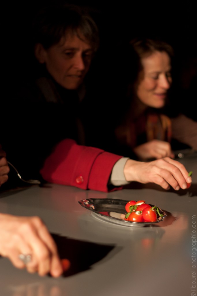 Une assiette sur une table avec des tomates. On voit à côtés de l'assiette derrière la table deux personnes assise. Dont une comme en train de prendre quelque chose.