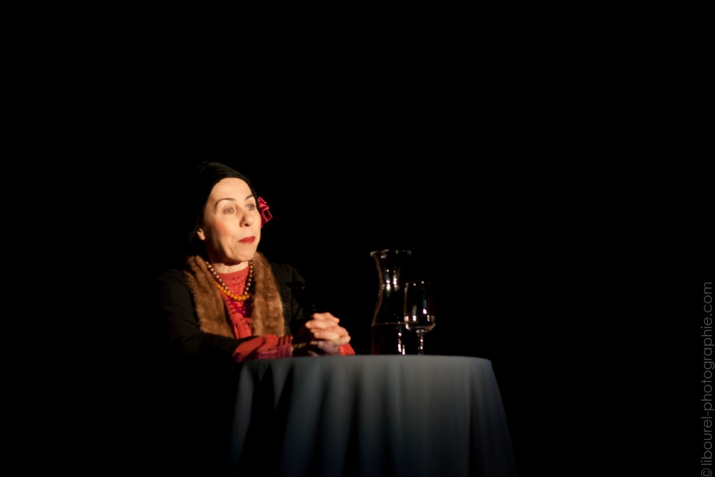 Une femme assise à une table. On dirait la table d'un restaurant. Elle porte un manteau à fourrure, un chapeau et un pull rouge.