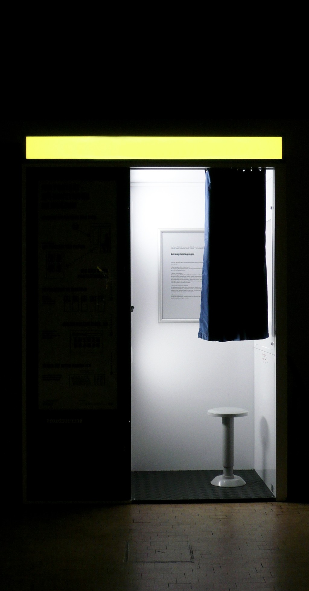 C'est l'image d'un photomaton dans le noir. Il y a de la lumière dans la cabine on aperçoit un petit rideau et un petit tabouret.