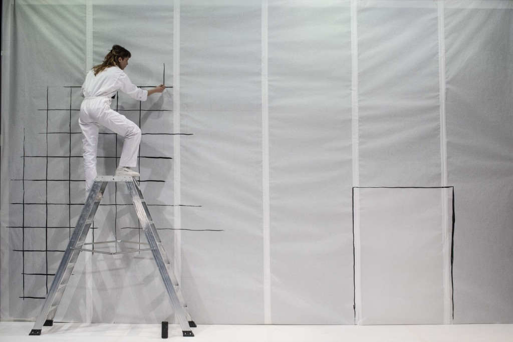 Une personne sur une échelle habillée avec une combinaison blanche, elle est en train de dessiner de quadrillage sur un mur, pas directement sur le mus mais sur le tissu ou la matiere qui récouvre le mur.