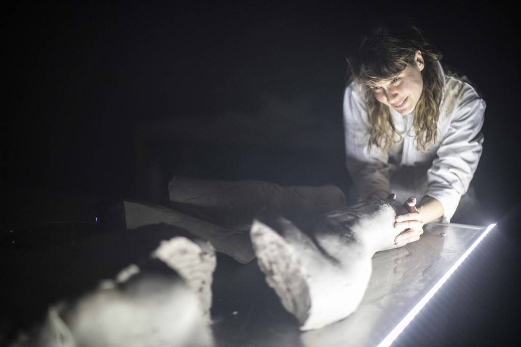 Une personne habillée en blanc sourie et touche des morceaux de statue sur une table illuminé au led.