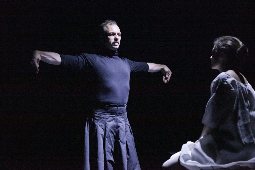 A gauche une personne en train de danser completement habillée en noir. A droite une personne assise habillée en blanc. On ne voit pas son visage, la photo est trop sombre.