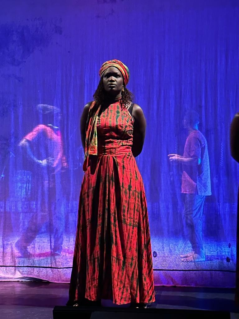Une personne se tient debout devant un rideau transparent. Derriere ce rideau deux personnes qui bougent. La personne devant le rideau porte une robe rouge et un turbant tipiques africans.