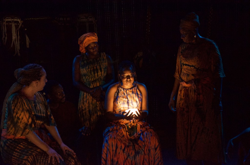 Quatre personnes dans la penombre. En semi cercle. Une personne est assise au centre et elle tient entre ses mains comme une petite lampe qui lui illumine le visage. On peut à peine distinguer les silhouettes et les visages des autres personnes.