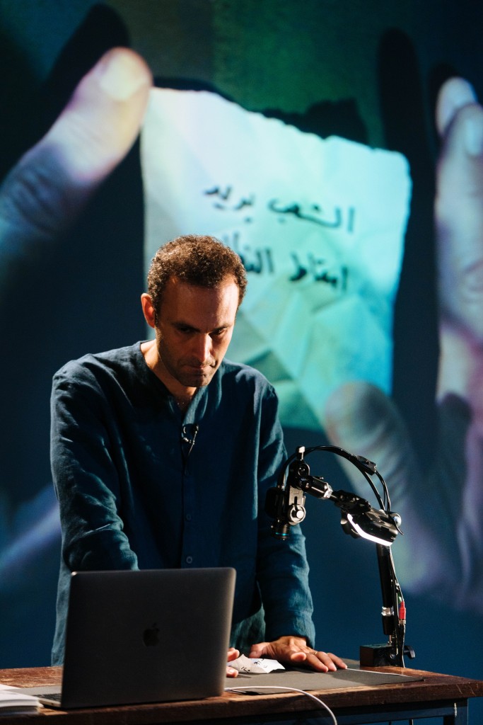 Une personne debout derrière un ordinateur sur un bureau. Derrière lui une projection vidéo : deux mains et une petite feuille de papier avec des écritures en arabe. La feuille c'est comme si elle avait été pliée.