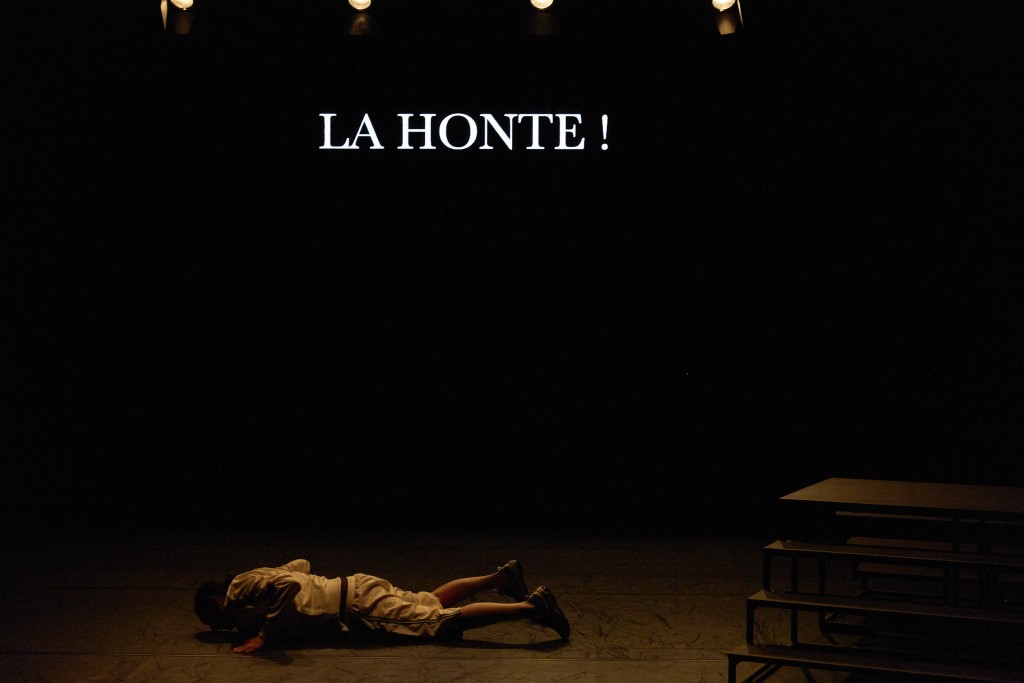Sur le fond de scène le mot LA HONTE ! est projeté en gros lettres. Léa Pohlhammer et couchée par terre le visage au sol.