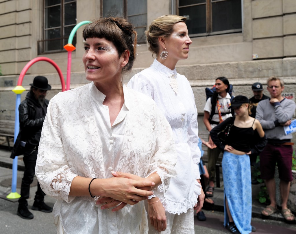 Dans cette image deux personnes habillées en blanc. Une des deux est très souriante. sur le fond des arches colorés et des personnes en train de regarder.