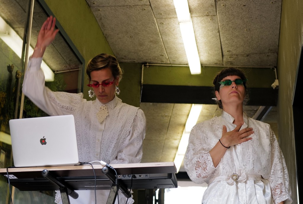 Deux personnes habillées en blanc en train de danser. La personne à gauche est derrier un ordi comme si elle mettait de la musique.