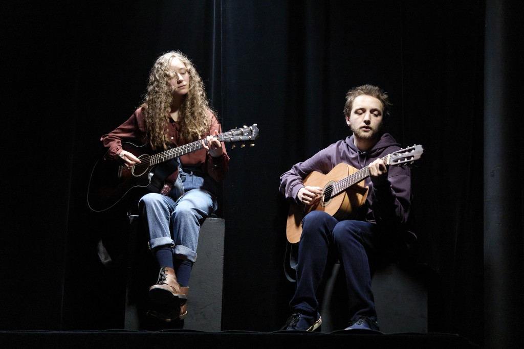 ce sont les musiciens du spectacle à gauche Tess Giordano, à droite Léon Boesch. Tess a les cheveux longs bouclés clair et elle porte une veste bordeau. Léon porte un pull violet et des jeans. les deux jouent de la guitarre