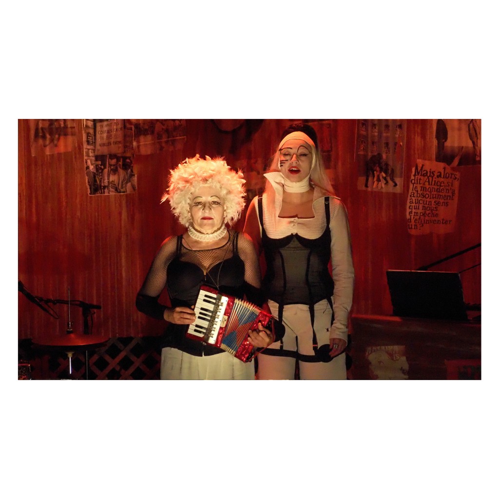 Une personne en train de jouer un petit accordeon. Elle porte une perruque balnche frisée. En haut en corset noir, derrière elle une autre personne avec une perruque blanche, chemise blanche, par dessus corset noir.