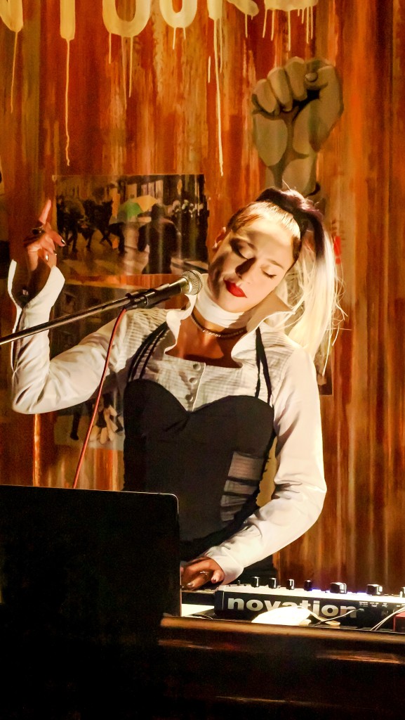 Une personne derrière une table de mixage, elle porte une chemise blanche et un corset noir. Elle porte une perruque blonde et un rouge à lèvre rouge.