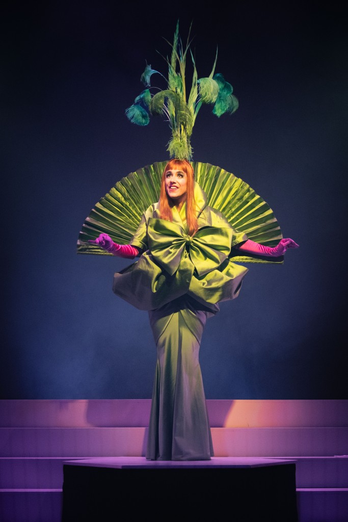 Une femme habillée en vert. Son costume évoque une plante verte au vue de son chapeau feuillou. Elle a des gants fuchsia, une perruque à cheveux longs et rouges.